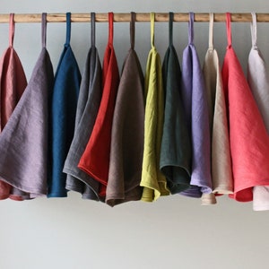 May include: A collection of 12 linen tea towels hanging on a wooden rod. The towels are in various colors, including pink, purple, blue, gray, brown, red, green, yellow, beige, and coral.