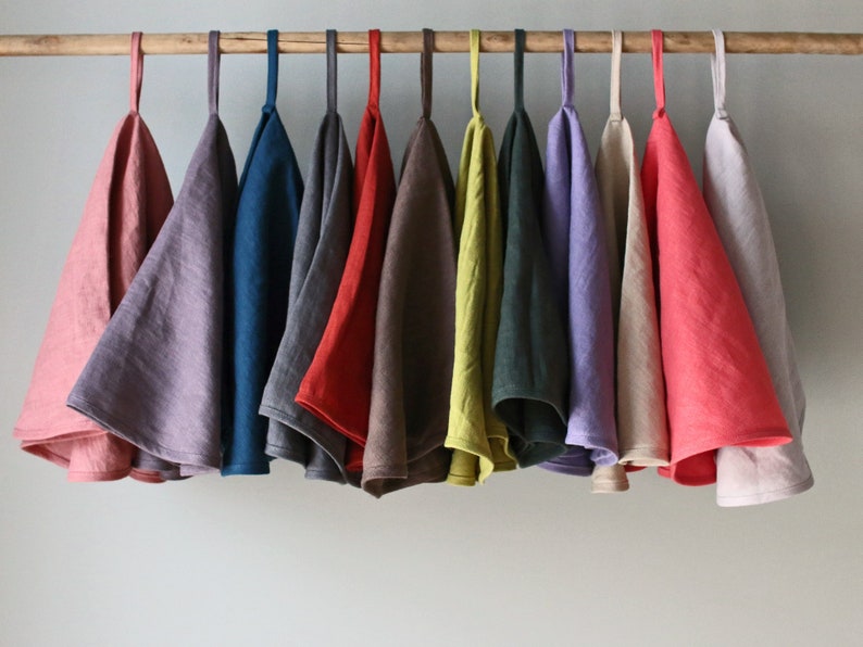 May include: A collection of 12 linen tea towels hanging on a wooden rod. The towels are in various colors, including pink, purple, blue, gray, brown, red, green, yellow, beige, and coral.