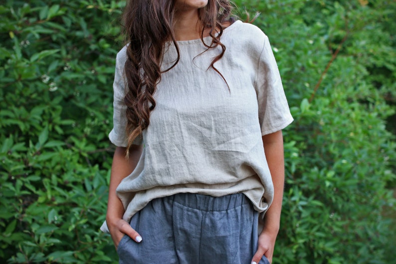 May include: A woman wearing a loose-fitting, light beige linen top with short sleeves and a pair of dark gray linen pants.