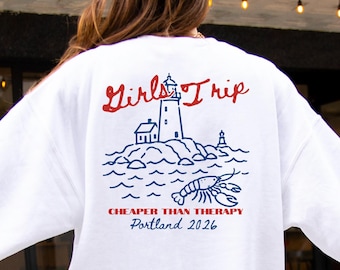 May include: A white long-sleeve sweatshirt featuring a back graphic with a lighthouse, ocean waves, and a lobster. The text reads Girls Trip in red, Cheaper Than Therapy in red, and Portland 2026 in blue. The design uses red and blue line art.
