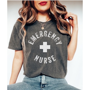Peut inclure: Un t-shirt gris foncé avec un motif blanc qui dit "Emergency Nurse" avec un signe plus blanc au centre.