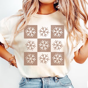 May include: A white t-shirt with a brown and white snowflake pattern. The shirt is a relaxed fit and has a crew neck. The text "IVORY" is visible in the bottom right corner of the image.