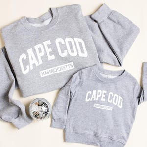 May include: Two gray sweatshirts with the text "CAPE COD MASSACHUSETTS" in white. One is adult-sized, the other is smaller. A silver disco ball sits near the sweatshirts. The sweatshirts are made of a soft material.