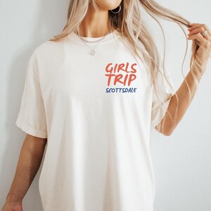 May include: Cream-colored t-shirt with the words "GIRLS TRIP" in red and "SCOTTSDALE" in blue. The shirt has short sleeves and a relaxed fit. The text is centered on the chest.