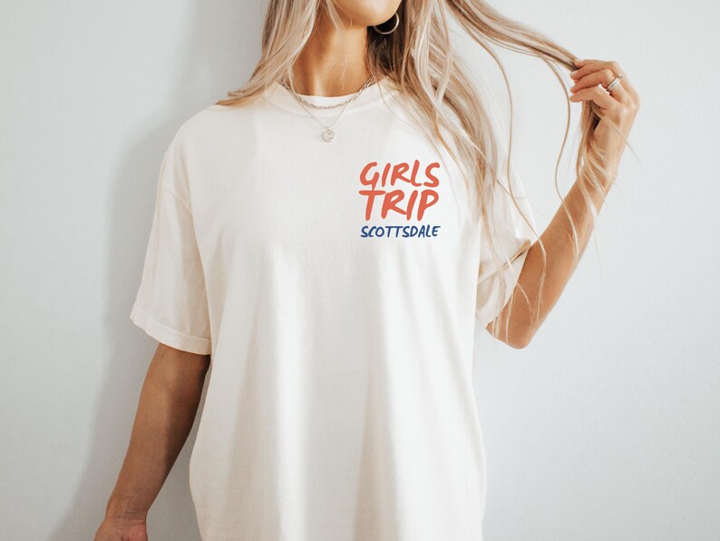May include: Cream-colored t-shirt with the words "GIRLS TRIP" in red and "SCOTTSDALE" in blue. The shirt has short sleeves and a relaxed fit. The text is centered on the chest.