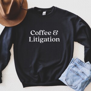 Op de afbeelding: Zwart sweatshirt met witte tekst die "Coffee & Litigation" zegt. Het sweatshirt ligt plat op een wit oppervlak met een bruine hoed en een blauwe jeans.