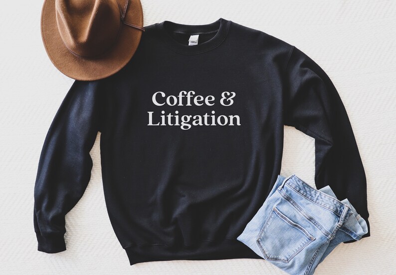 Op de afbeelding: Zwart sweatshirt met witte tekst die "Coffee & Litigation" zegt. Het sweatshirt ligt plat op een wit oppervlak met een bruine hoed en een blauwe jeans.