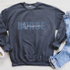 K&ouml;nnte beinhalten: Ein dunkelgraues Sweatshirt mit den W&ouml;rtern "NURSE trauma" in blauen und wei&szlig;en Buchstaben. Das Sweatshirt wird mit einer blauen Jeans und wei&szlig;en Turnschuhen kombiniert. Das Bild ist auf einer wei&szlig;en strukturierten Oberfl&auml;che aufgenommen.