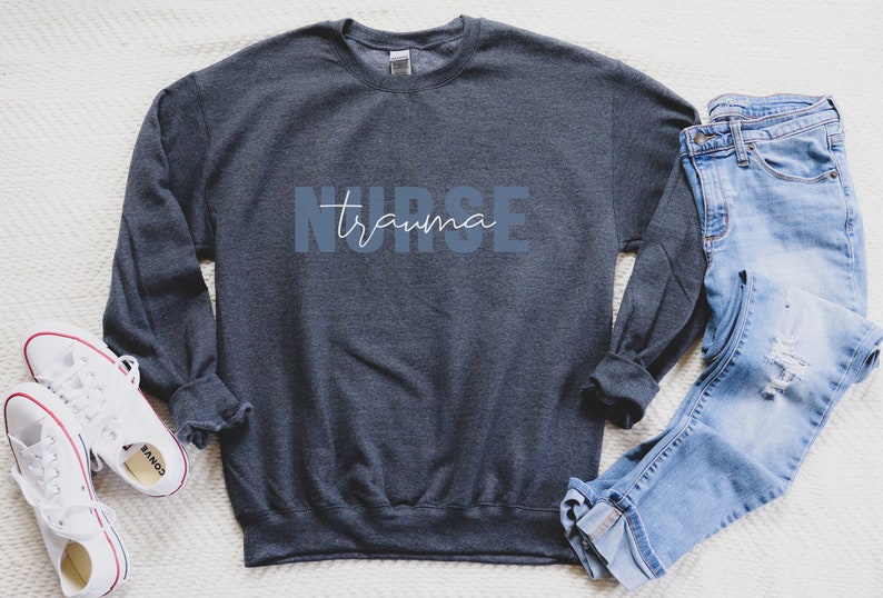 K&ouml;nnte beinhalten: Ein dunkelgraues Sweatshirt mit den W&ouml;rtern "NURSE trauma" in blauen und wei&szlig;en Buchstaben. Das Sweatshirt wird mit einer blauen Jeans und wei&szlig;en Turnschuhen kombiniert. Das Bild ist auf einer wei&szlig;en strukturierten Oberfl&auml;che aufgenommen.