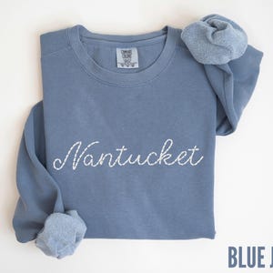 May include: A blue jean-colored sweatshirt with the embroidered word "Nantucket" in white script. The long-sleeved shirt is folded, with the sleeves and a scrunchie visible. The text "BLUE JEAN" is at the bottom right.