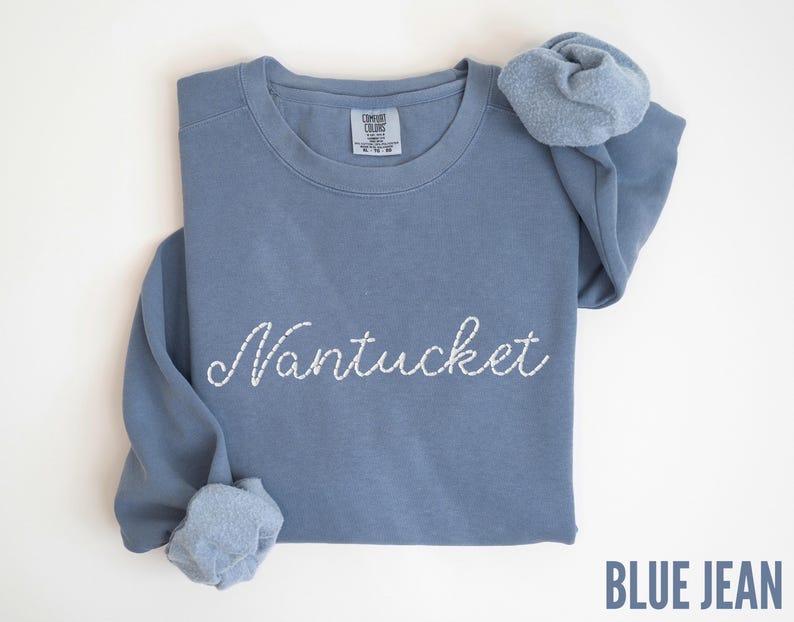 May include: A blue jean-colored sweatshirt with the embroidered word "Nantucket" in white script. The long-sleeved shirt is folded, with the sleeves and a scrunchie visible. The text "BLUE JEAN" is at the bottom right.