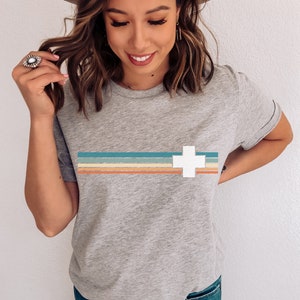 Puede incluir: Una camiseta gris con una cruz blanca y rayas de colores a trav&eacute;s del pecho. La camiseta est&aacute; hecha de un material suave y c&oacute;modo.