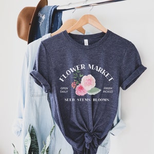 May include: A navy blue t-shirt with a white graphic that says "Flower Market" with a floral design and the text "Open Daily, Fresh Picked, Seed, Stems, Blooms."