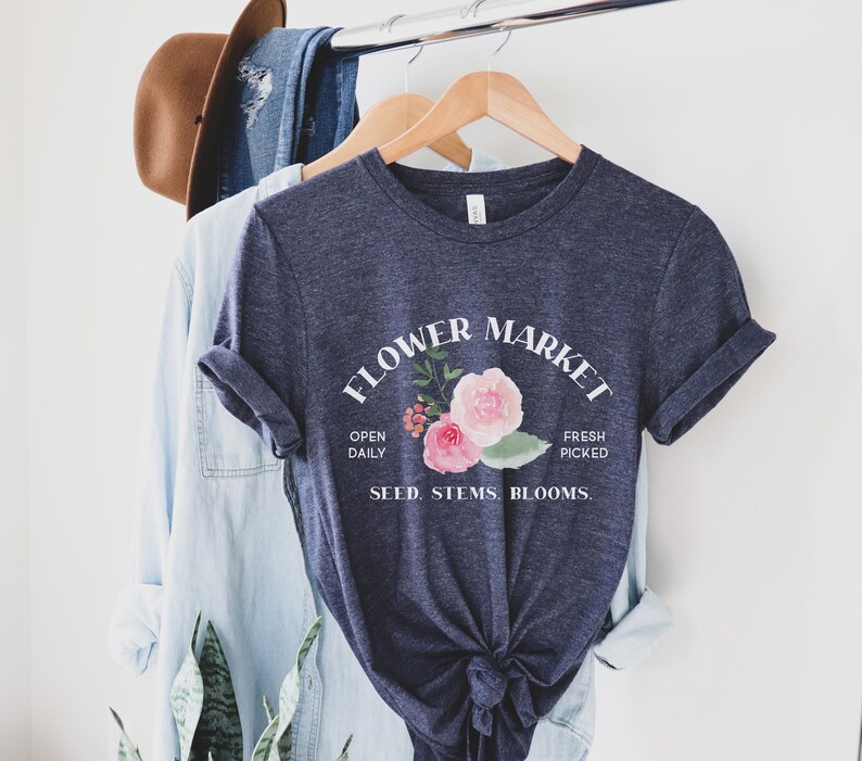 May include: A navy blue t-shirt with a white graphic that says "Flower Market" with a floral design and the text "Open Daily, Fresh Picked, Seed, Stems, Blooms."