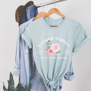 May include: A light blue t-shirt with a white graphic that says "Flower Market" with a floral design and the text "Open Daily, Fresh Picked, Seed, Stems, Blooms."