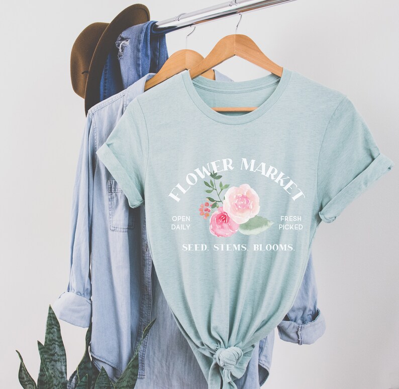 May include: A light blue t-shirt with a white graphic that says "Flower Market" with a floral design and the text "Open Daily, Fresh Picked, Seed, Stems, Blooms."