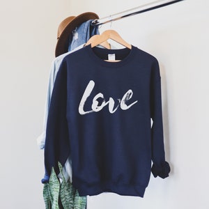 Puede incluir: Una sudadera azul marino con un dise&ntilde;o de pincelada blanca que dice "Love" en la parte delantera.
