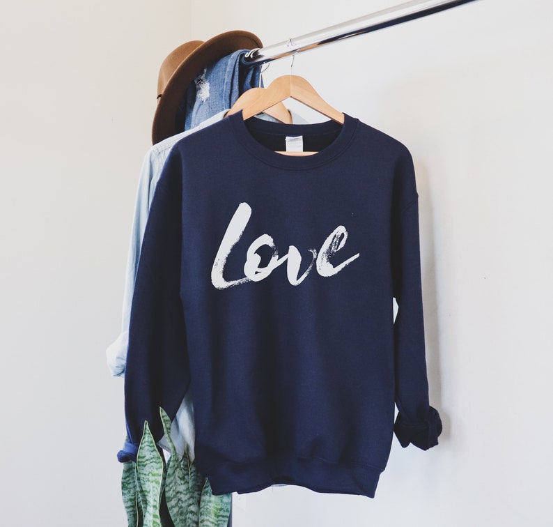 Puede incluir: Una sudadera azul marino con un dise&ntilde;o de pincelada blanca que dice "Love" en la parte delantera.