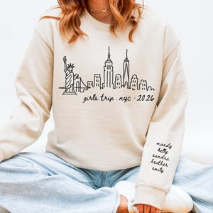 May include: Beige sweatshirt with a black line drawing of the New York City skyline, including the Statue of Liberty. The text "girls trip . nyc . 2026" is below the skyline. The right sleeve has the names "mandy, kelly, sandra, heather, emily".