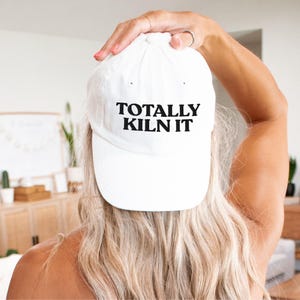May include: White baseball cap with the text "TOTALLY KILN IT" in black block letters. The hat is worn backward. The image is taken from behind a person with blonde hair.