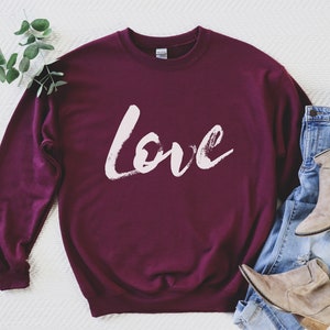 Puede incluir: Sudadera color burdeos con un dise&ntilde;o de pinceladas blancas que dice "love" en la parte delantera.