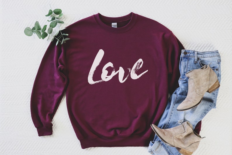 Puede incluir: Sudadera color burdeos con un dise&ntilde;o de pinceladas blancas que dice "love" en la parte delantera.