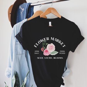 May include: Black t-shirt with a white graphic that says "Flower Market" with a floral design and the text "Open Daily, Fresh Picked, Seed. Stems. Blooms."