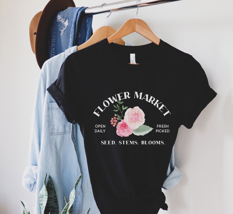 May include: Black t-shirt with a white graphic that says "Flower Market" with a floral design and the text "Open Daily, Fresh Picked, Seed. Stems. Blooms."