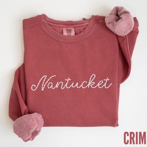 May include: Crimson-colored sweatshirt with the word "Nantucket" embroidered in white script across the front. The long sleeves have a rolled-up cuff. The sweatshirt is made of a soft material.