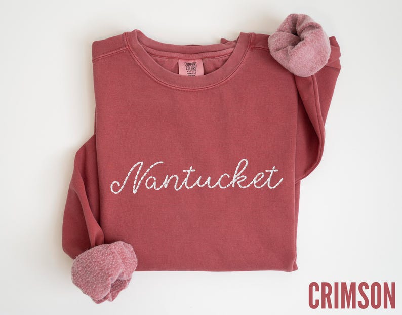 May include: Crimson-colored sweatshirt with the word "Nantucket" embroidered in white script across the front. The long sleeves have a rolled-up cuff. The sweatshirt is made of a soft material.