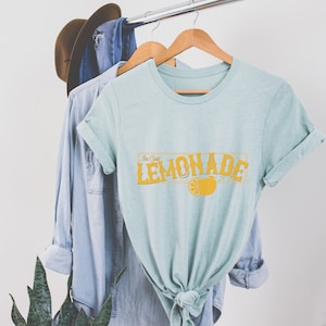 K&ouml;nnte beinhalten: Ein hellblaues T-Shirt mit einer gelben Grafik, auf der "Ice Cold Lemonade" mit einer Zitronenscheibe steht.