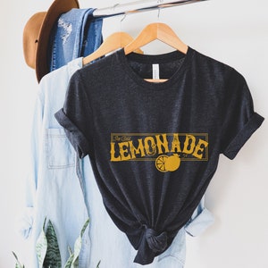 K&ouml;nnte beinhalten: Ein dunkelgraues T-Shirt mit einer gelben Grafik, auf der "Ice Cold Lemonade" mit einer Zitronenscheibe steht.