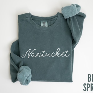 May include: A blue spruce sweatshirt with the word "Nantucket" embroidered in white script. The sleeves are cuffed, and the text "BLUE SPRUCE" is printed in a sans-serif font in the lower right corner. The sweatshirt is laid flat on a white surface.