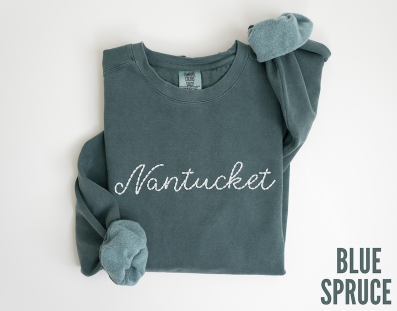 May include: A blue spruce sweatshirt with the word "Nantucket" embroidered in white script. The sleeves are cuffed, and the text "BLUE SPRUCE" is printed in a sans-serif font in the lower right corner. The sweatshirt is laid flat on a white surface.