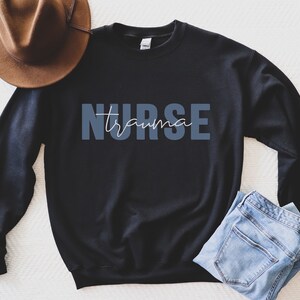 K&ouml;nnte beinhalten: Schwarzes Sweatshirt mit dem Text "NURSE trauma" in Blau und Wei&szlig;. Ein brauner Hut und blaue Jeans sind ebenfalls im Bild. Das Sweatshirt hat einen Rundhalsausschnitt.