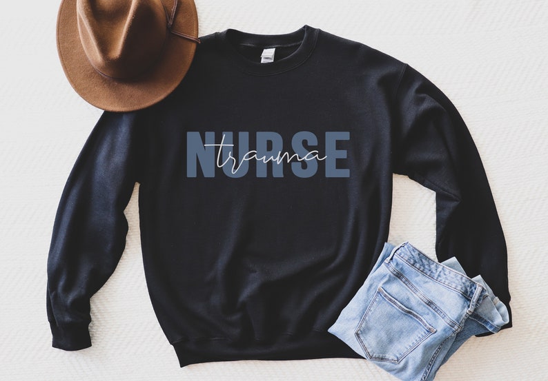 K&ouml;nnte beinhalten: Schwarzes Sweatshirt mit dem Text "NURSE trauma" in Blau und Wei&szlig;. Ein brauner Hut und blaue Jeans sind ebenfalls im Bild. Das Sweatshirt hat einen Rundhalsausschnitt.