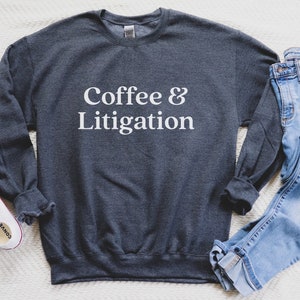 Op de afbeelding: Een donkergrijs sweatshirt met de tekst "Coffee & Litigation" in witte letters. Het sweatshirt ligt plat op een wit oppervlak met een blauwe jeans en witte sneakers.