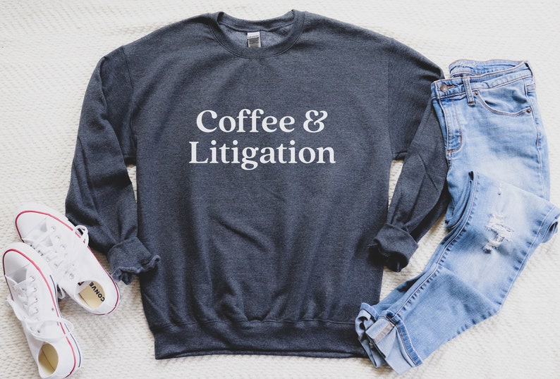 Op de afbeelding: Een donkergrijs sweatshirt met de tekst "Coffee & Litigation" in witte letters. Het sweatshirt ligt plat op een wit oppervlak met een blauwe jeans en witte sneakers.