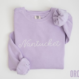 May include: A lavender-colored sweatshirt with the word "Nantucket" embroidered in white script. The sweatshirt has a crew neck and long sleeves with cuffed wrists. The word "ORCHID" is printed in the bottom right corner.