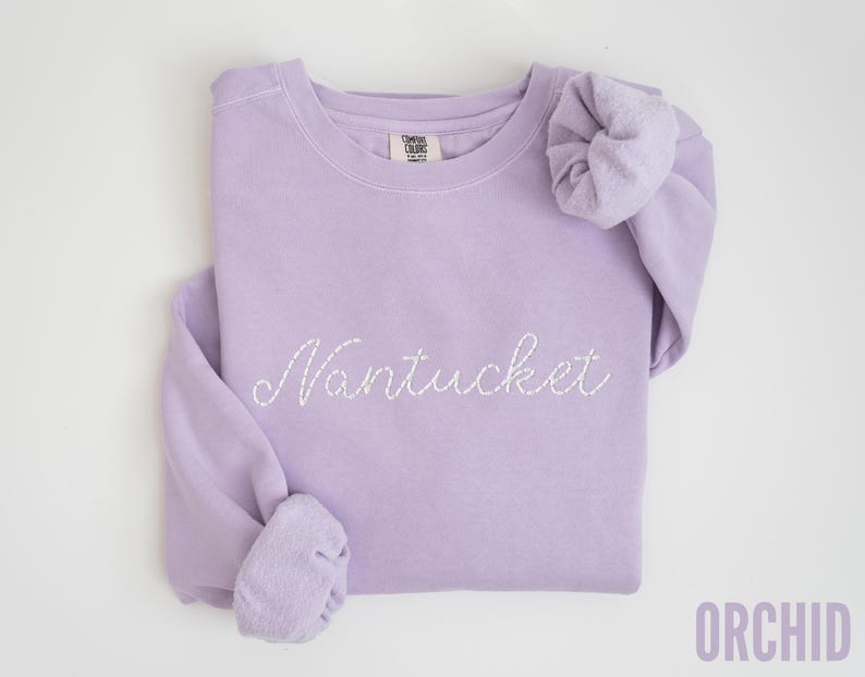 May include: A lavender-colored sweatshirt with the word "Nantucket" embroidered in white script. The sweatshirt has a crew neck and long sleeves with cuffed wrists. The word "ORCHID" is printed in the bottom right corner.