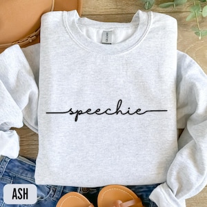 May include: A light gray sweatshirt with the word "speechie" in black script. The sweatshirt is folded on top of blue jeans and sandals. The word "ASH" is in a white box in the bottom left corner.