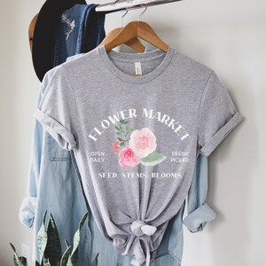 May include: Gray t-shirt with a white graphic that says "Flower Market" with a floral design and the text "Open Daily, Fresh Picked, Seed, Stems, Blooms."