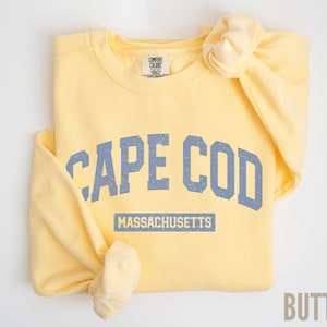 May include: A light yellow sweatshirt with the words "CAPE COD" in blue block letters and "MASSACHUSETTS" underneath. The sweatshirt is folded with the sleeves tied in a knot. The word "BUTTER" is in the bottom right corner.