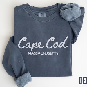 May include: A denim blue sweatshirt with the words "Cape Cod Massachusetts" in white script. The sweatshirt is folded with the sleeves rolled up, and the word "DENIM" is printed in a darker blue font.