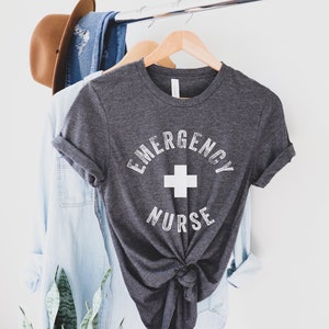 May include: A dark gray t-shirt with a white graphic that says "Emergency Nurse" with a white plus sign in the center.