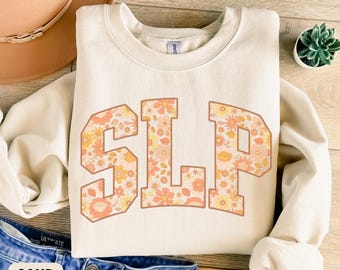 May include: Cream-colored sweatshirt with the letters "SLP" in a floral pattern. The letters are outlined in a brown color. The sweatshirt is folded on a wooden surface with a pair of blue jeans and a hat.