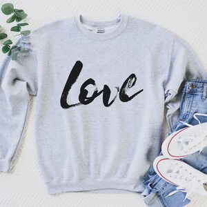 Puede incluir: Una sudadera gris con la palabra "love" escrita en negro con pinceladas. La sudadera est&aacute; colocada sobre una superficie blanca con un par de zapatillas blancas y unos vaqueros azules.