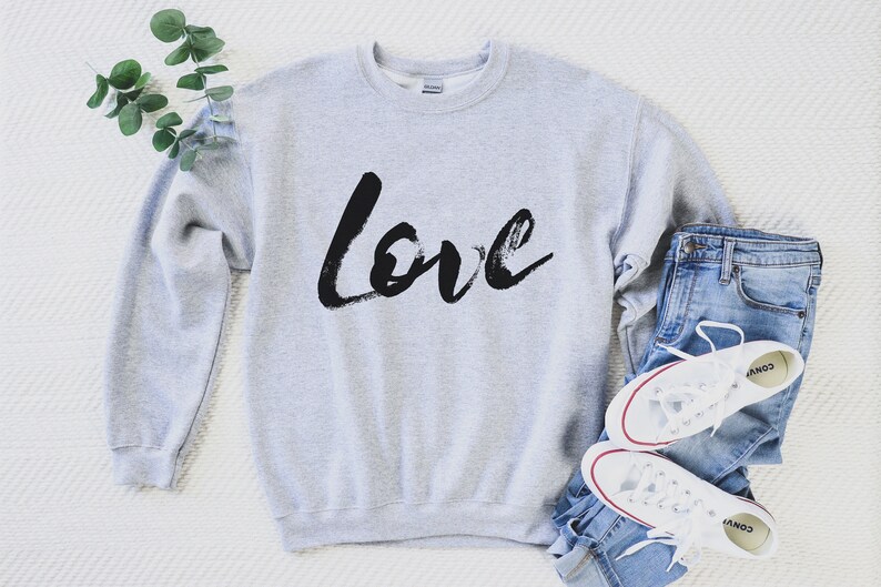 Puede incluir: Una sudadera gris con la palabra "love" escrita en negro con pinceladas. La sudadera est&aacute; colocada sobre una superficie blanca con un par de zapatillas blancas y unos vaqueros azules.