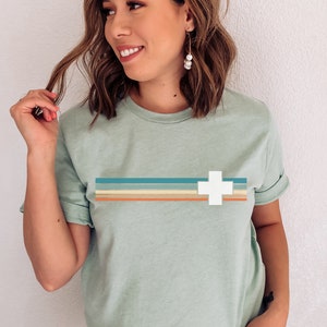 Puede incluir: Una camiseta verde claro con un dise&ntilde;o retro que presenta una cruz blanca y rayas horizontales en tonos azules, naranjas y marrones.