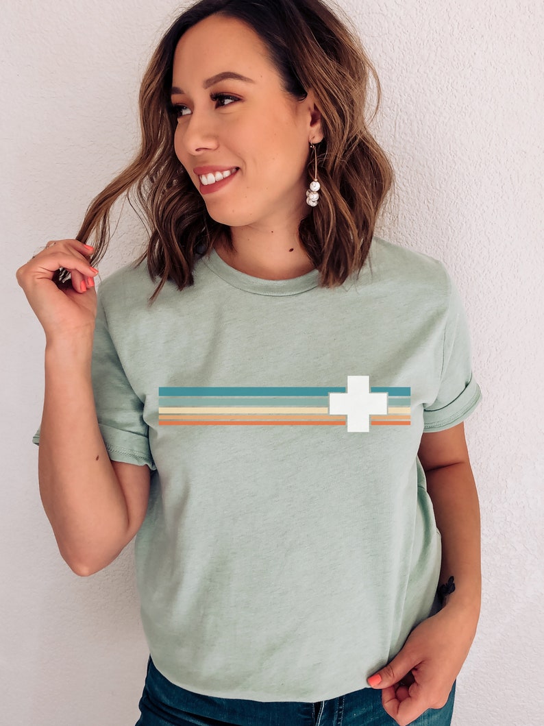 Puede incluir: Una camiseta verde claro con un dise&ntilde;o retro que presenta una cruz blanca y rayas horizontales en tonos azules, naranjas y marrones.
